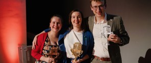 Drie mensen poseren op een podium; twee houden microfoons vast, terwijl de derde een shirt met een bedrukte microfoonafbeelding draagt.