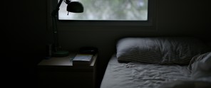 Sfeervolle slaapkamer met een onopgemaakt bed, een nachtkastje met een boek en een groene lamp, en natuurlijk licht dat door het raam naar binnen valt.