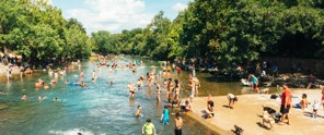 Drukke rivier met zwemmers en zonaanbidders, omringd door bomen onder een heldere blauwe lucht.