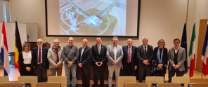 A group of ten people in formal attire stands in front of a presentation, with an aerial view of a building and agricultural land in the background.