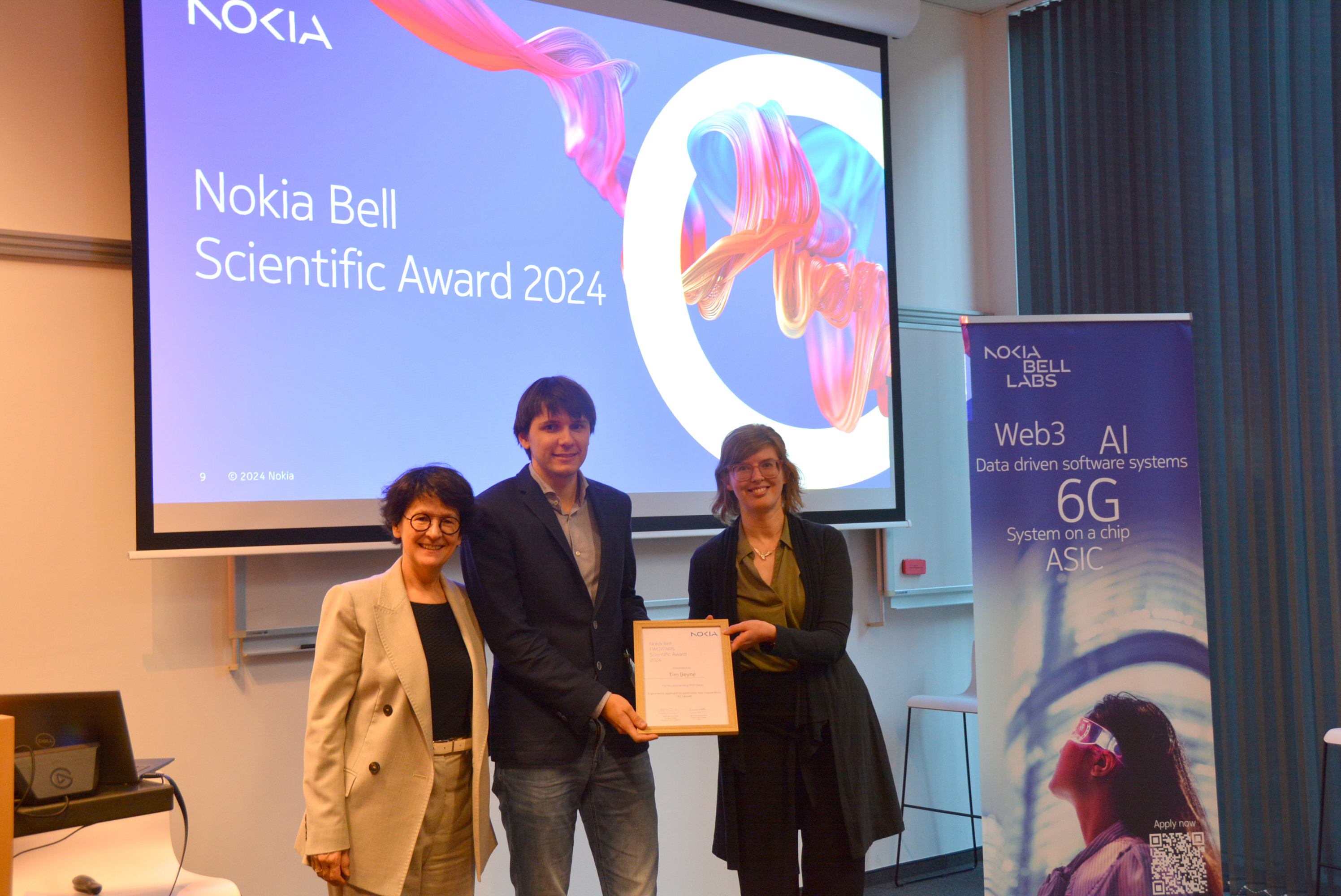 Drie mensen staan op het podium en tonen een certificaat, met een achtergrondbanner die technologie benadrukt.